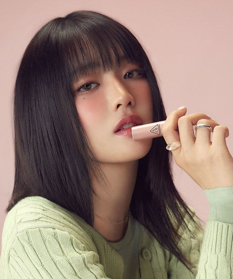 Woman wearing soft makeup holding 3CE Glaze Lip Glow 3.2g against a pastel pink background.