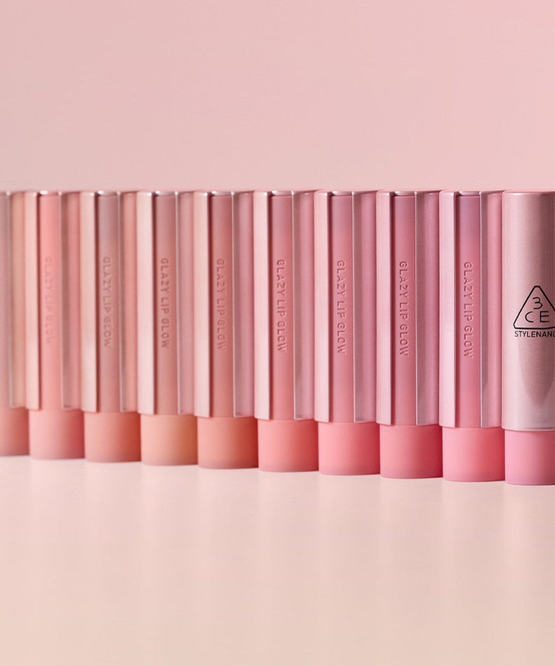 Assorted shades of 3CE Glaze Lip Glow 3.2g lipsticks lined up on a light pink background.
