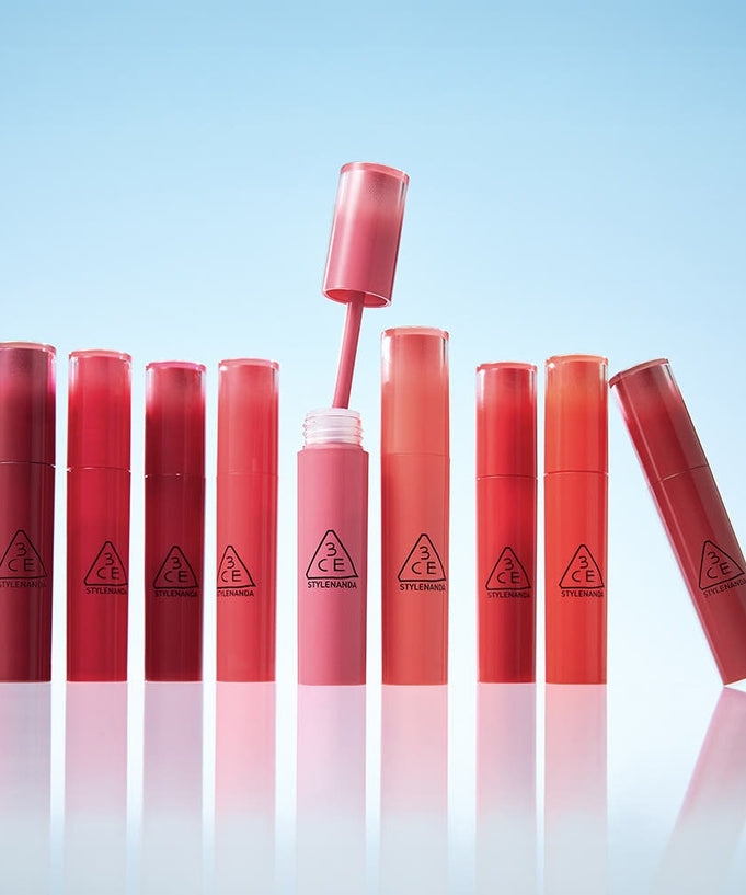 Eight tubes of 3CE Lazy Pop Lip Stain arranged on a reflective surface against a blue background, showcasing vibrant and diverse lip colors.