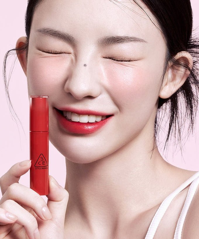 Woman smiling and holding 3CE Lazy Pop Lip Stain in red shade.