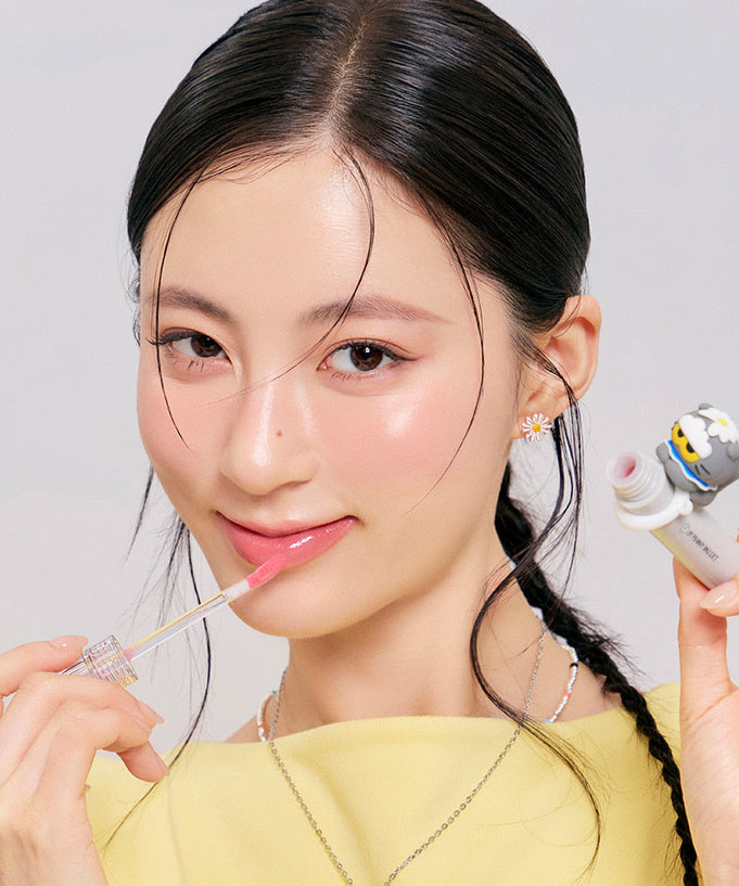 Woman applying ROMAND x Joanne Friends Glasting Color Gloss in a playful packaging with a smile, showcasing its glossy effect.