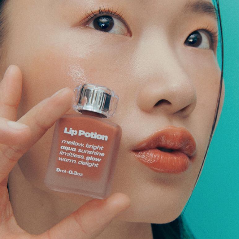 Close-up of a model holding ALTERNATIVE STEREO Lip Potion Aqua Glow Tint, showcasing glossy hydrated lips and product bottle.