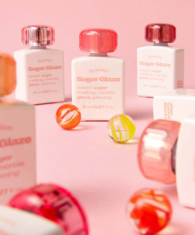 Display of ALTERNATIVE STEREO Lip Potion Sugar Glaze bottles with colorful design on a pink background, highlighting product aesthetics.