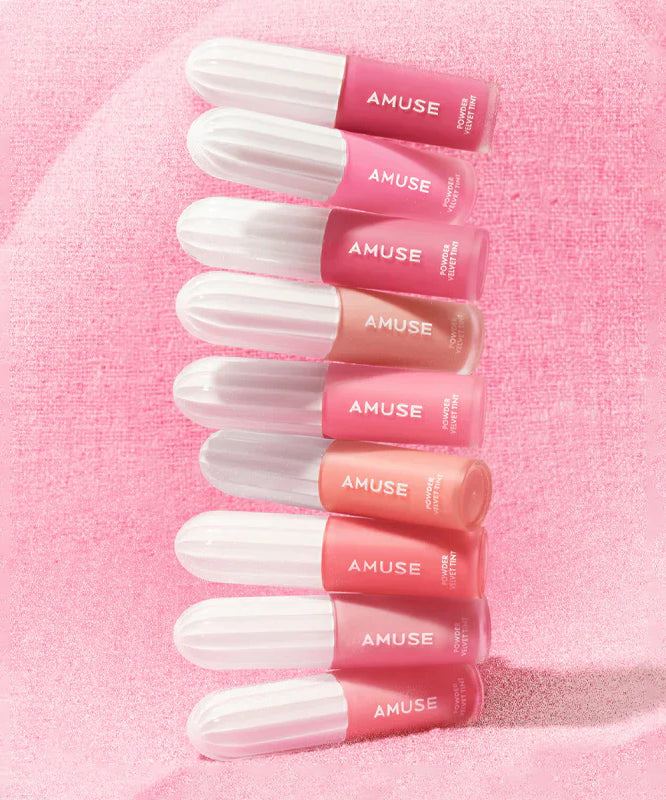 Various shades of AMUSE Powder Velvet Tint lip products on a soft pink background.