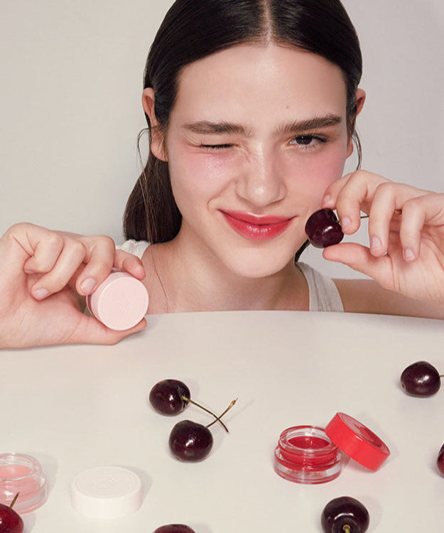 Woman holding ANILLO Mellow Glow Balm with cherries and balm containers on table.