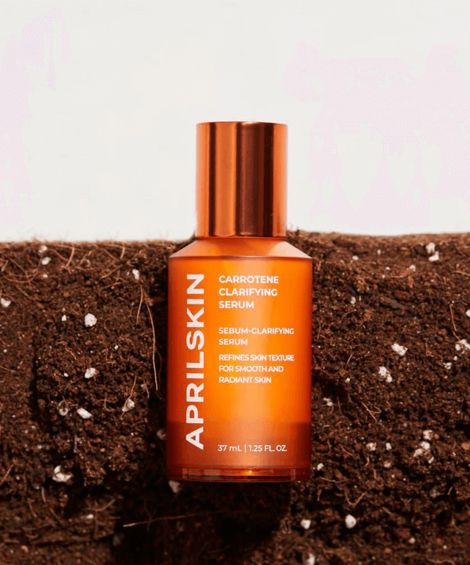 Orange bottle of APRILSKIN Carrotene Clarifying Serum on soil background for smooth and radiant skin.