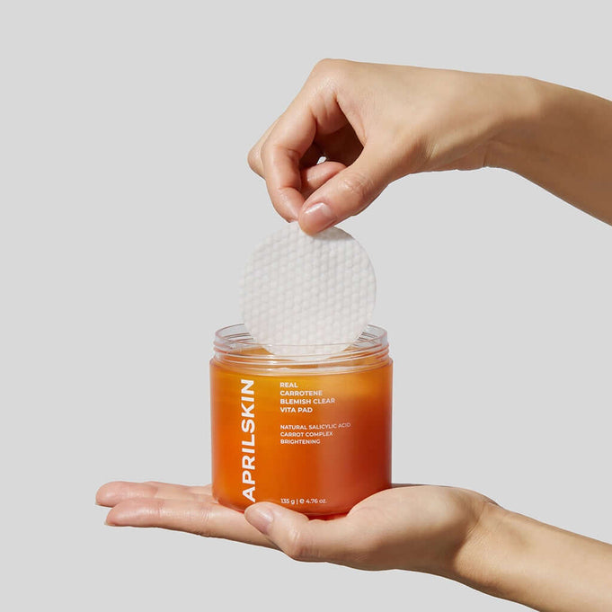 Hands holding APRILSKIN Carrotene Clarifying Vita Pad with visible cotton pad in orange jar