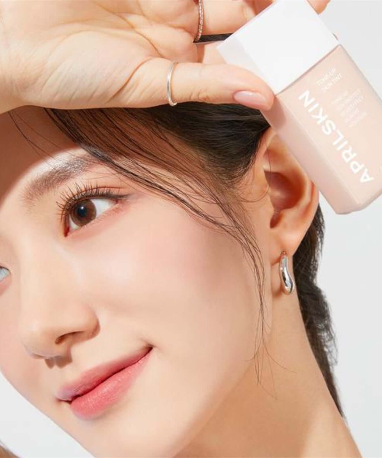 Woman holding APRILSKIN Tone-Up Skin Tint showcasing glowing skin and makeup application.