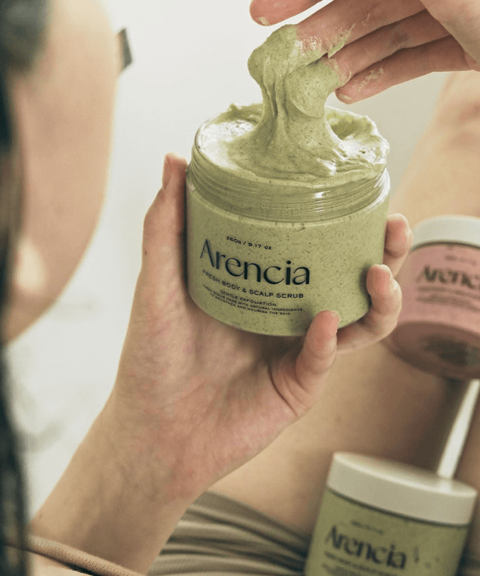Person using ARENCIA Fresh Cloud Body Scrub 260g, scooping creamy texture from jar, with pink and green containers in background.