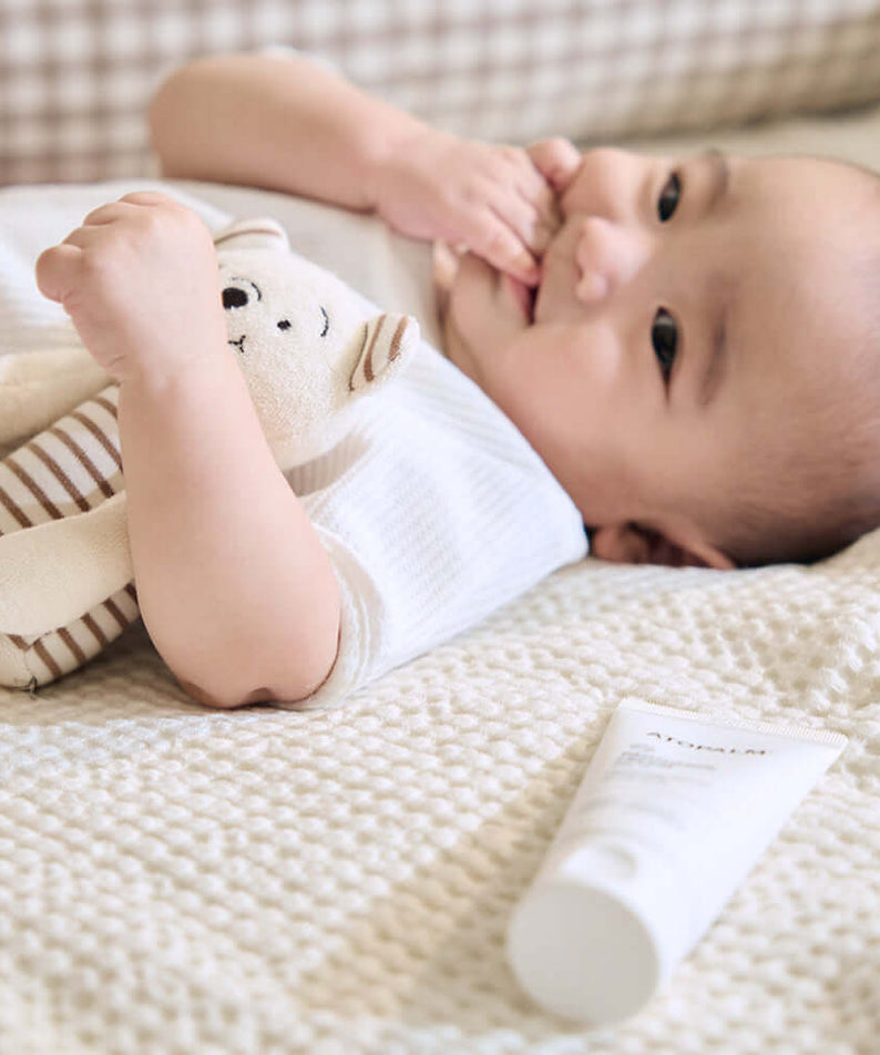 Baby lying on a blanket with stuffed toy next to ATOPALM Panthenol Lotion 180ml tube.