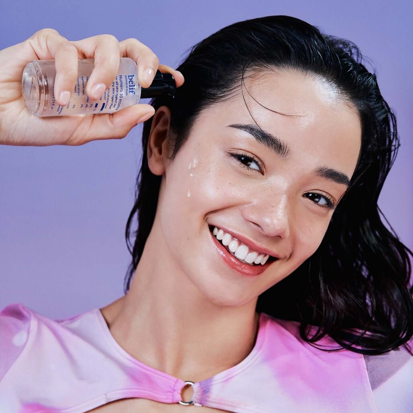 Woman applying BELIF Numero 10 Essence 75ml on her face, smiling brightly with a purple background.