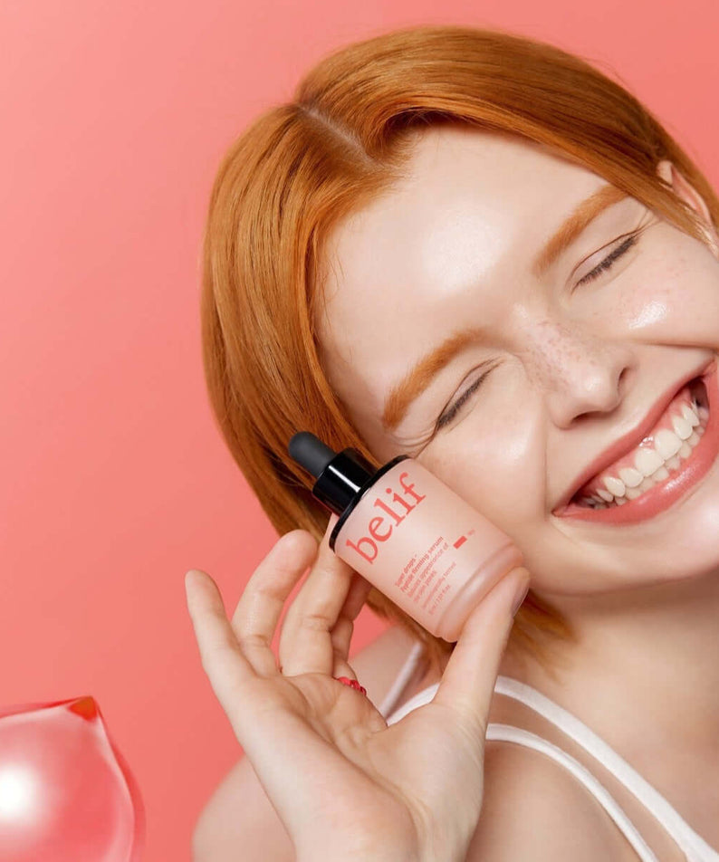 Smiling woman holding BELIF Super Drops Peptide Firming Serum 30ml against pink background.