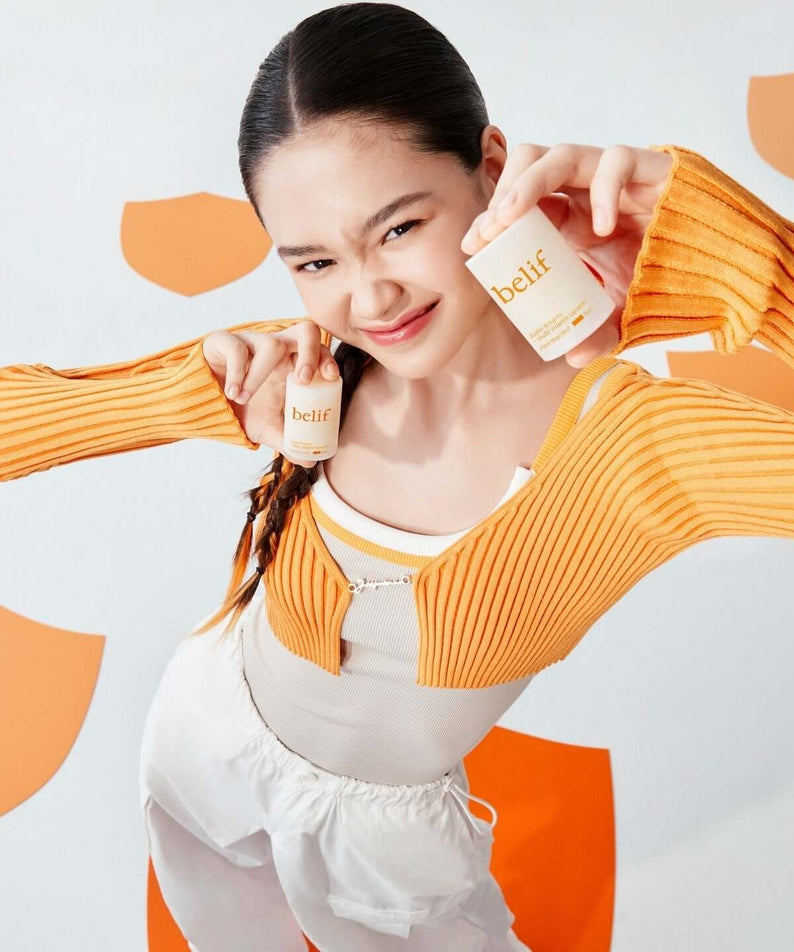 Person holding BELIF Super Knight Multi Vitamin Lipcerin products wearing orange sweater against orange and white background.