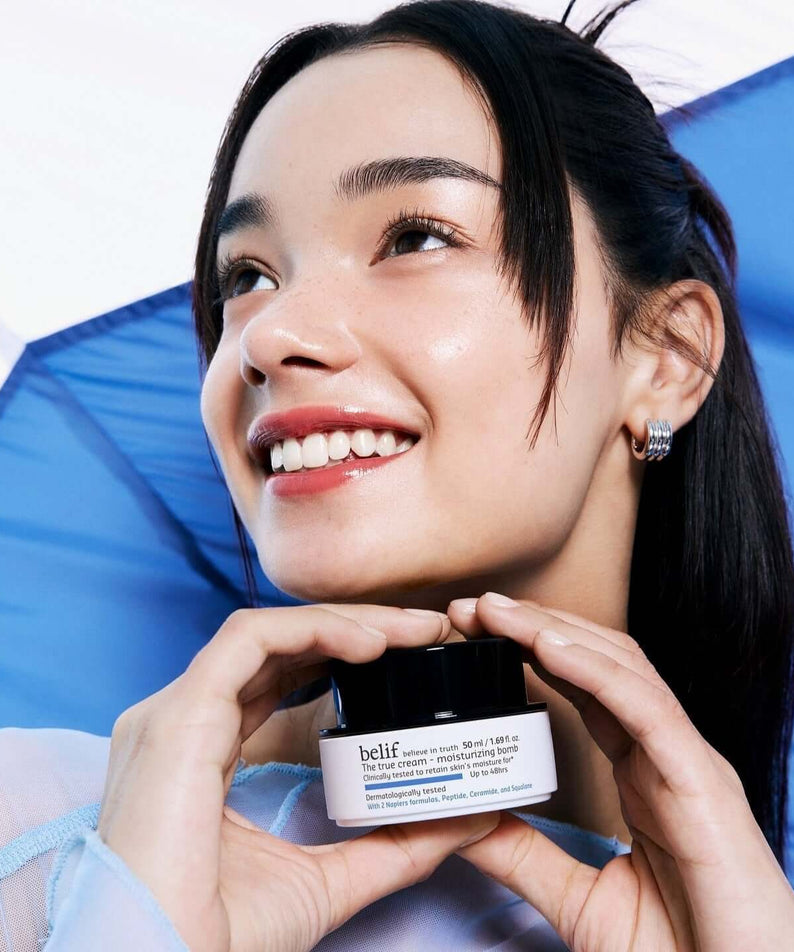 Smiling person holding BELIF The True Cream Moisturizing Bomb 50ml against a blue backdrop.