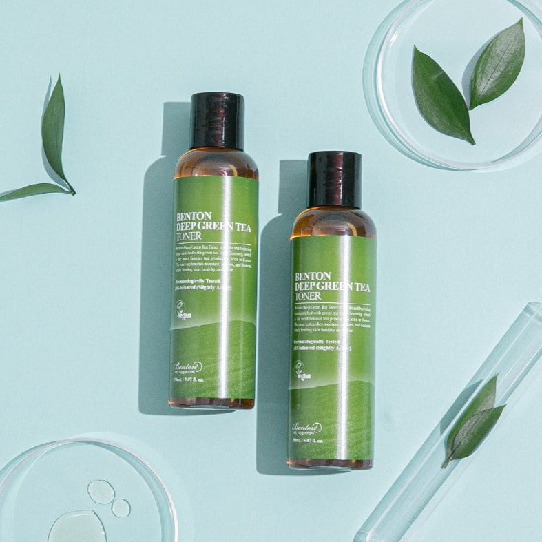 Two bottles of BENTON Deep Green Tea Toner 150ml with fresh green tea leaves on a blue background.
