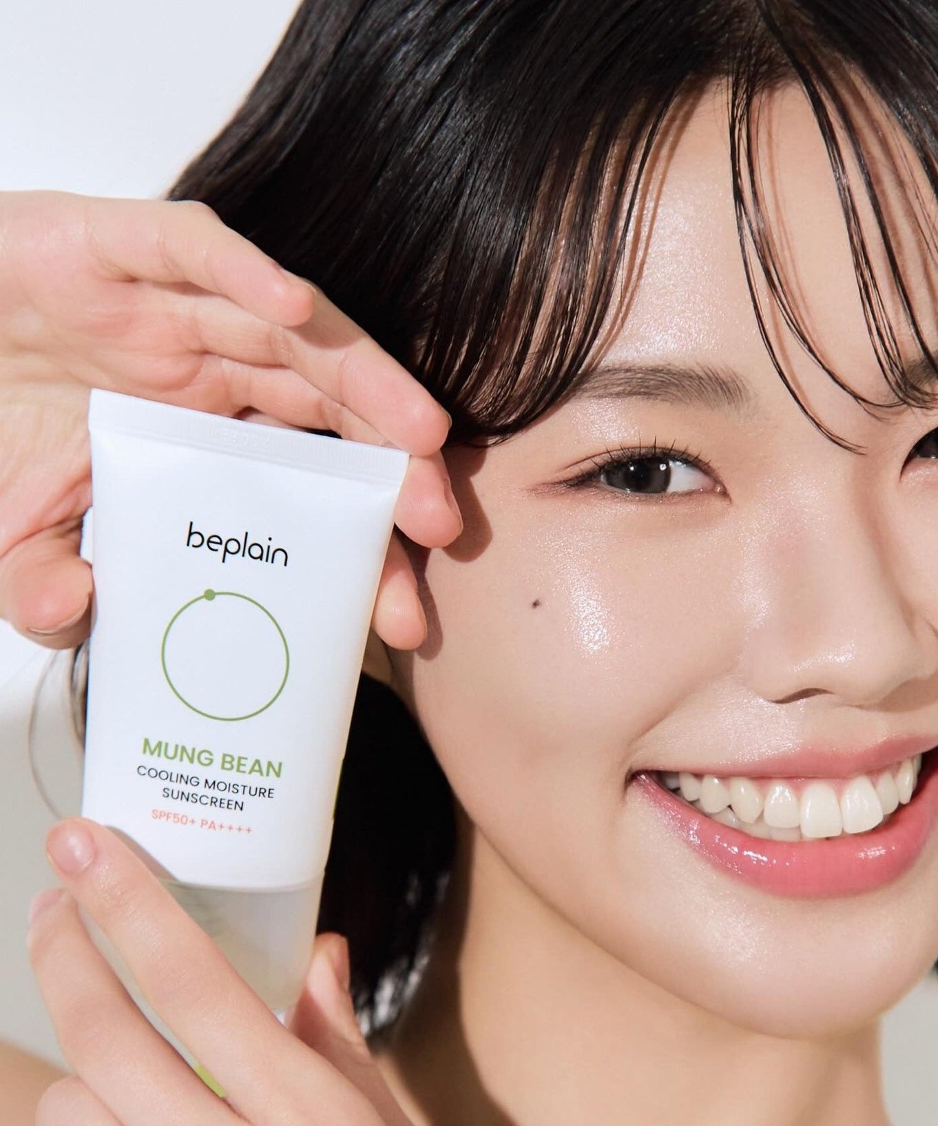 Smiling woman holding BEPLAIN Green Bean Cooling Moisture Sun Cream 50ml tube next to her face.