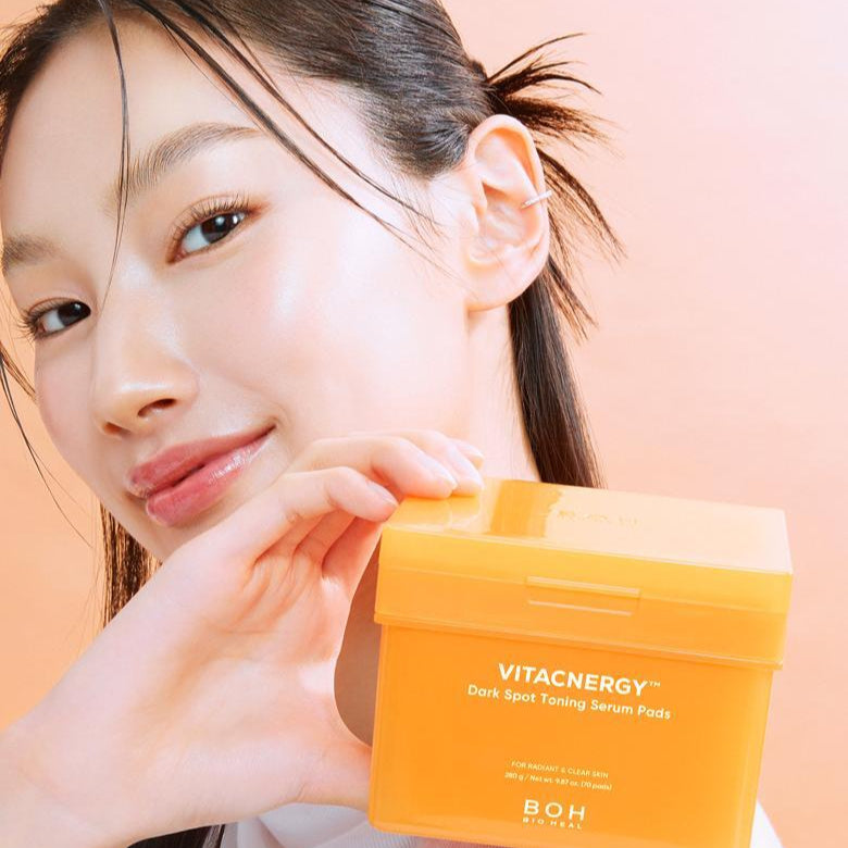 Smiling person holding a box of BIO HEAL BOH Vita Synergy Spot Toning Serum Pad 70 sheets against a peach background.