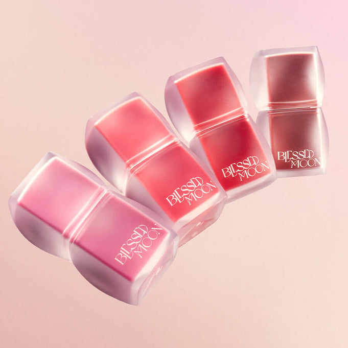 BLESSED MOON Veil Blur Lip Tint in various shades on a pink background, four lip tints lined up with sleek packaging.