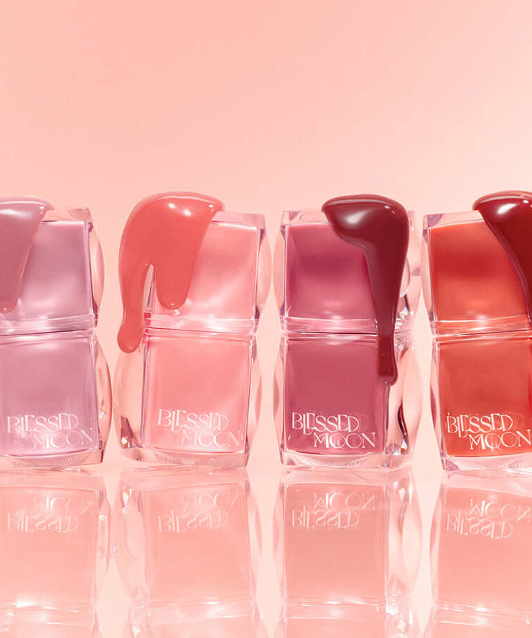 Assorted shades of BLESSED MOON Veil Glow Lip Tint in transparent containers against a pink background.