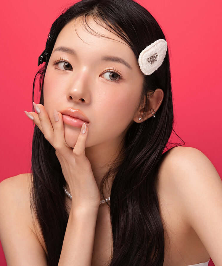 Model wearing BLESSED MOON Veil Glow Lip Tint with natural makeup and hair accessory against a pink background.