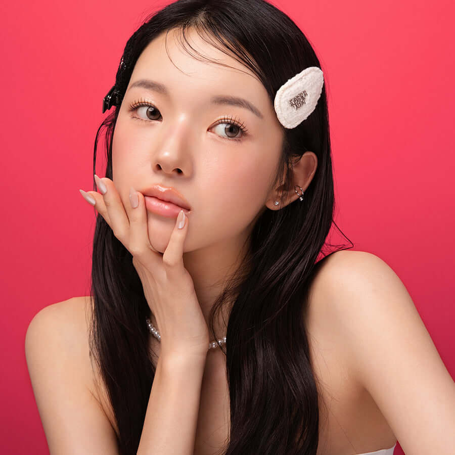 Model wearing BLESSED MOON Veil Glow Lip Tint with natural makeup and hair accessory against a pink background.