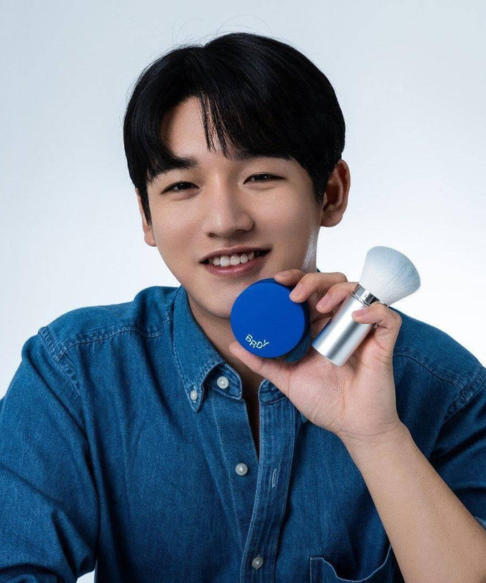 Smiling person holding B.READY Blue Sebum Powder and applicator brush, showcasing the product in a promotional setting.