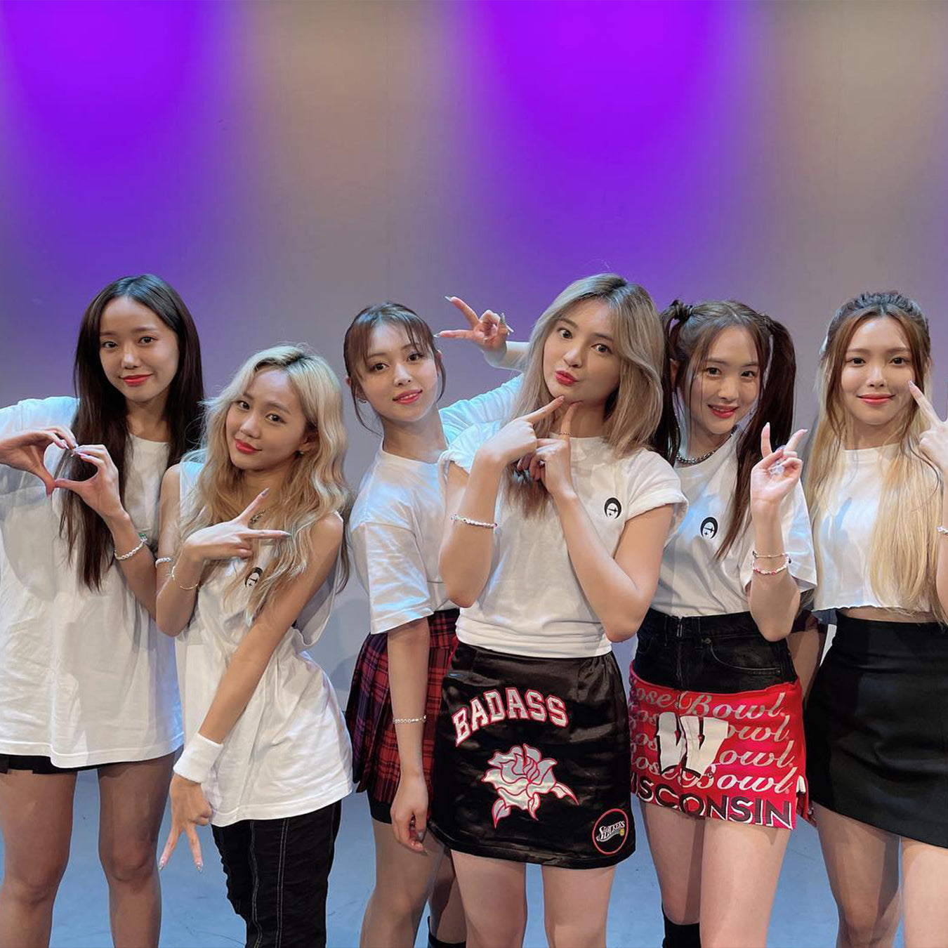 A group of six young women posing together on stage with a bright pink and purple backdrop, wearing casual and trendy fashion.