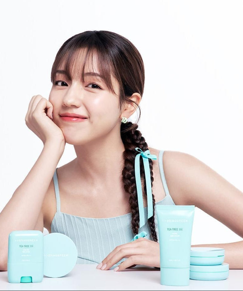 Smiling woman with BRINGGREEN Tea Tree Cica Tone Up Sun Cushion and skincare products on table.