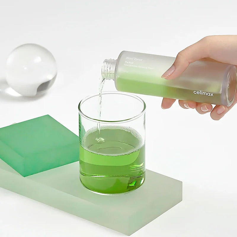 CELIMAX The Real Noni Moisture Balancing Toner 320ml being poured into a glass with green fluid, showcasing product texture.
