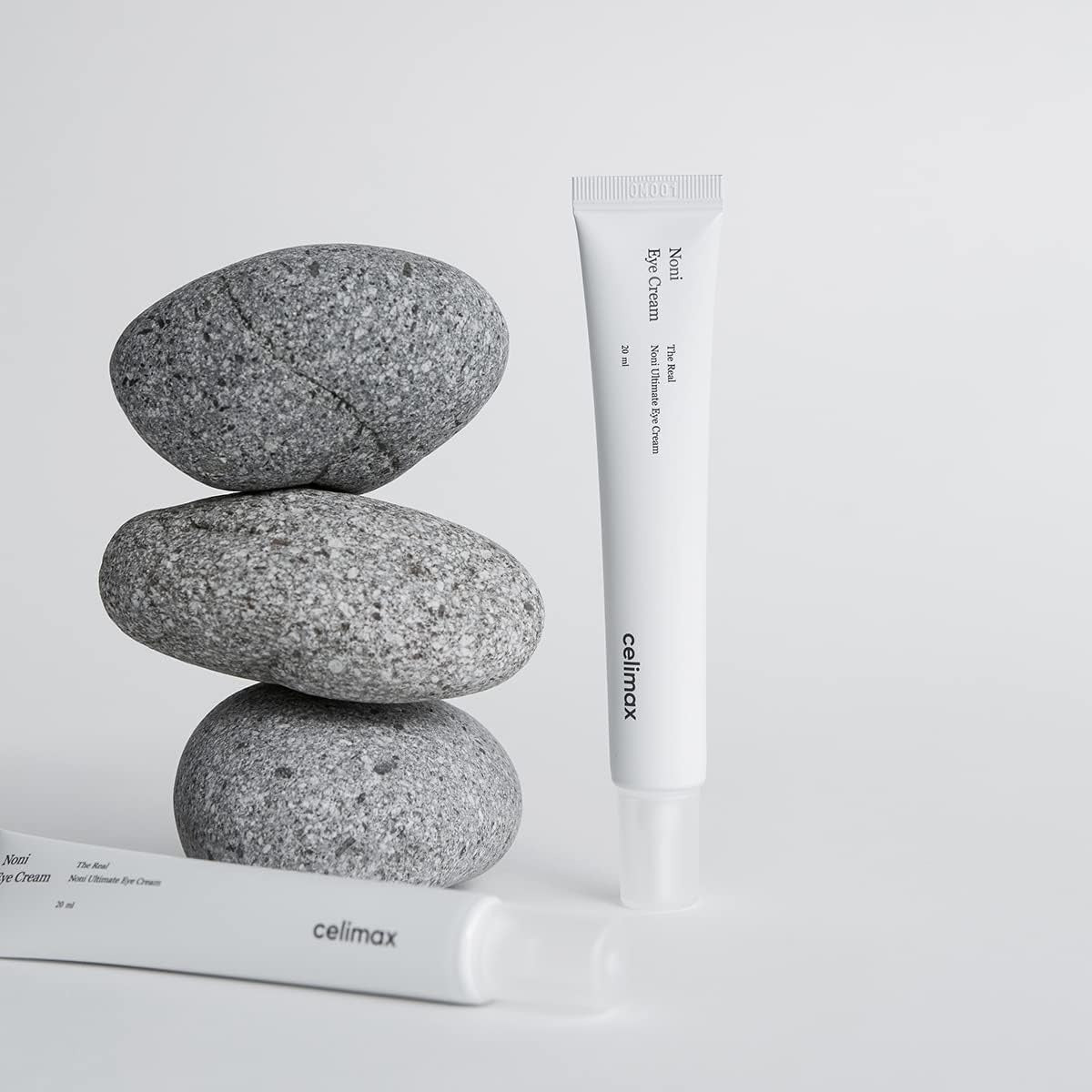 CELIMAX The Real Noni Ultimate Eye Cream 20ml tube displayed with stones for skincare product advertisement.