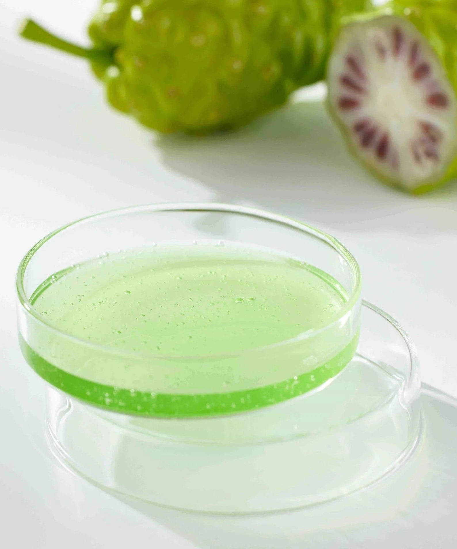 CELIMAX Noni Ampoule Calming+Radiance 100ml in a clear dish with sliced noni fruit in the background