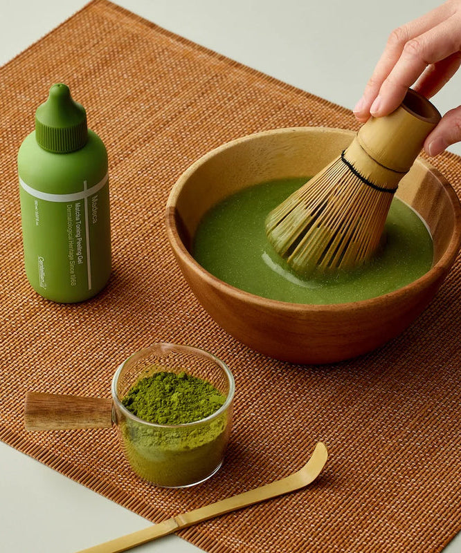 Matcha tea preparation with a bamboo whisk, cup of matcha powder, and a green bottle on a brown mat. Ideal for matcha enthusiasts.