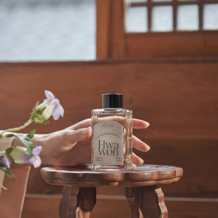 Hand holding CHWI (CHI) Hwa Won Diffuser 100ml bottle on a wooden table with flowers; premium fragrance for a calming ambiance.