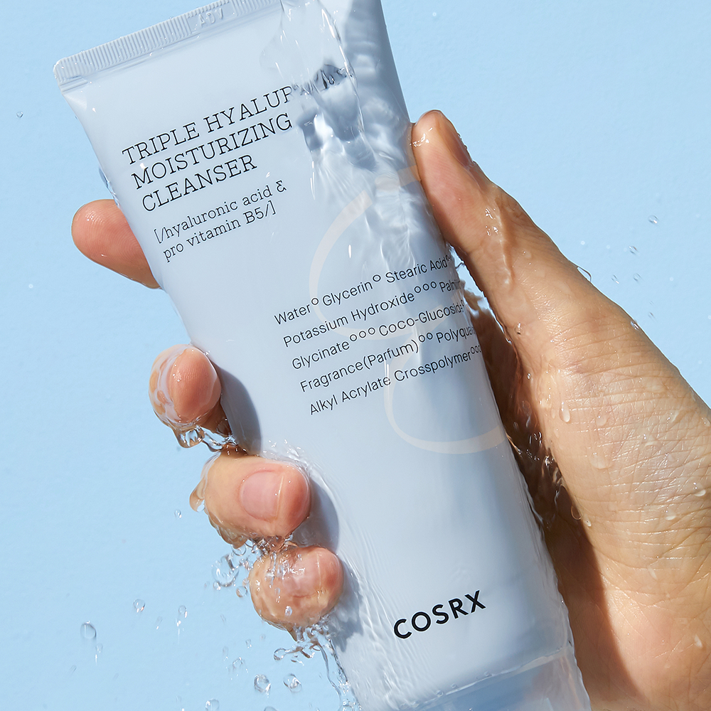Hand holding COSRX Hydrium triple Hyaluronic Moisturizing Cleanser 150ml with water splash, skincare product display.