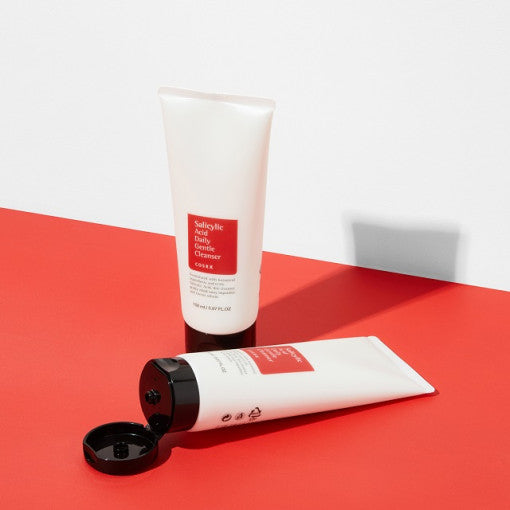 Two tubes of COSRX Salicylic Acid Daily Gentle Cleanser 150ml on a red surface, one standing and one lying down with open cap.