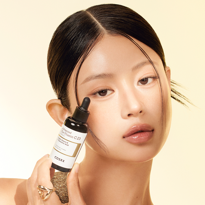 Woman holding COSRX Advanced The Vitamin C 23 Serum bottle against a neutral background.
