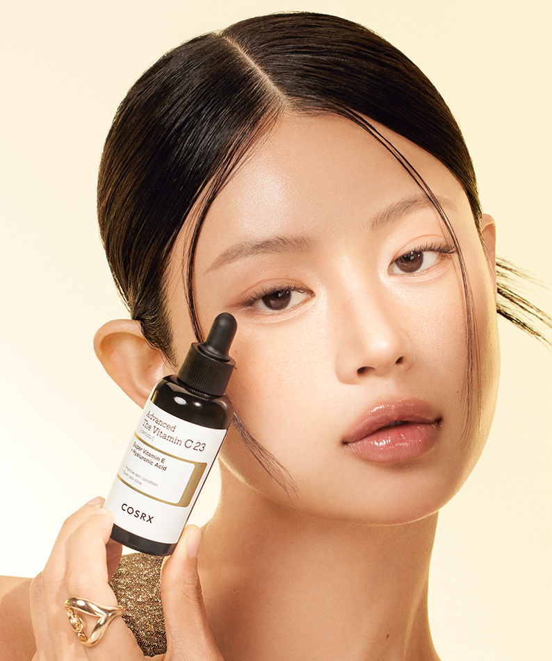 Woman holding COSRX Advanced The Vitamin C 23 Serum bottle against a neutral background.