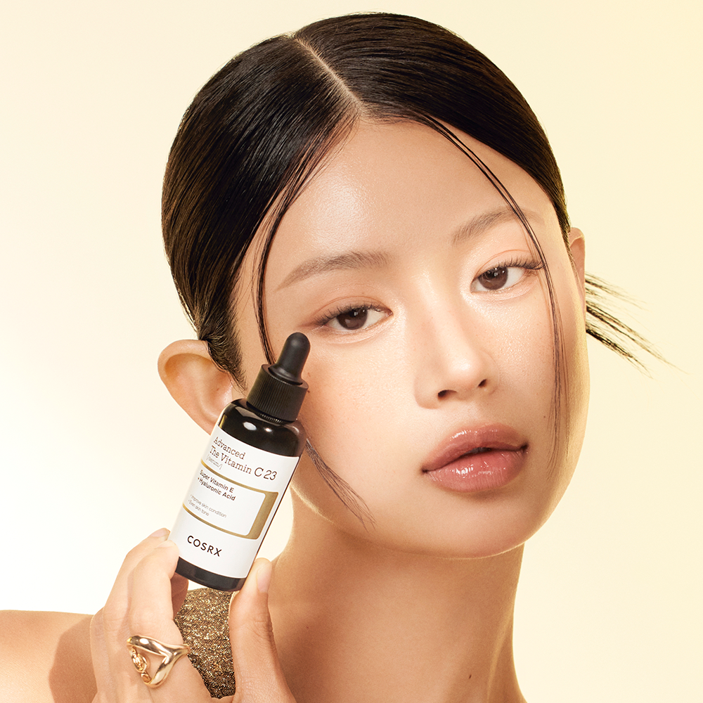Woman holding COSRX Advanced The Vitamin C 23 Serum bottle against a neutral background.