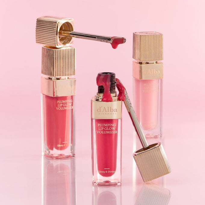 D'ALBA Plumbing Lip Glow Mood Volumizer in pink and red shades with gold accents, set against a soft pink background.