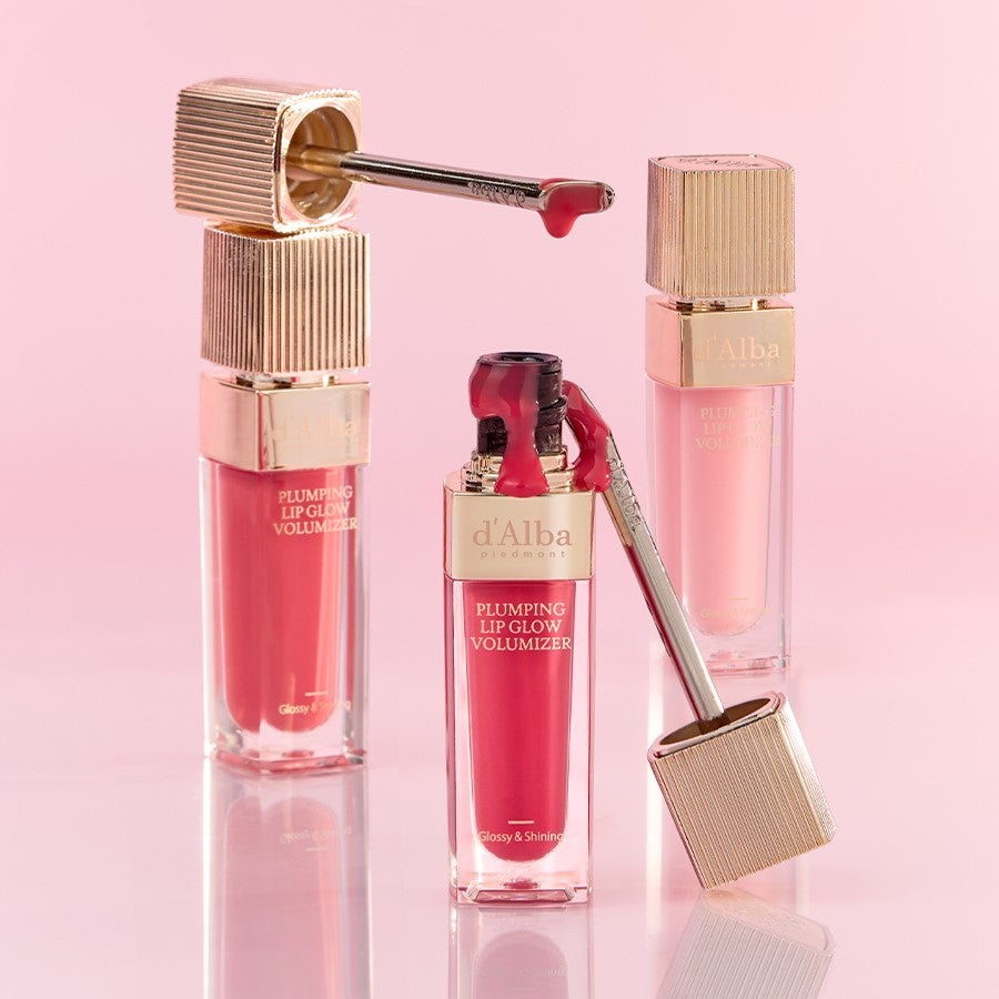 D'ALBA Plumbing Lip Glow Mood Volumizer in pink and red shades with gold accents, set against a soft pink background.