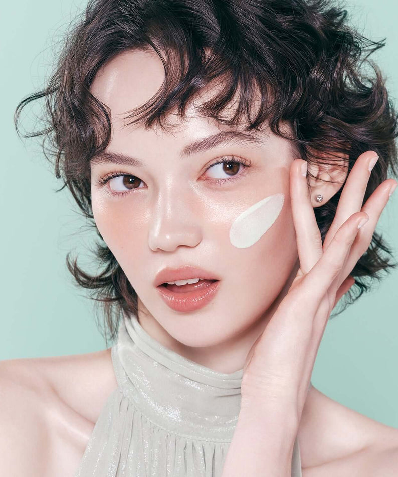 Woman applying DEAR DAHLIA Prime Layer Correcting Primer to her face against a soft green background.