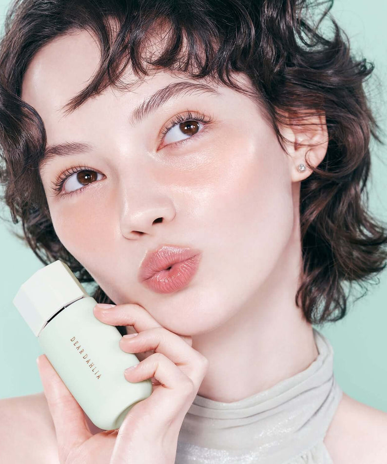 Person holding DEAR DAHLIA Prime Layer Correcting Primer with glowing skin, close-up.
