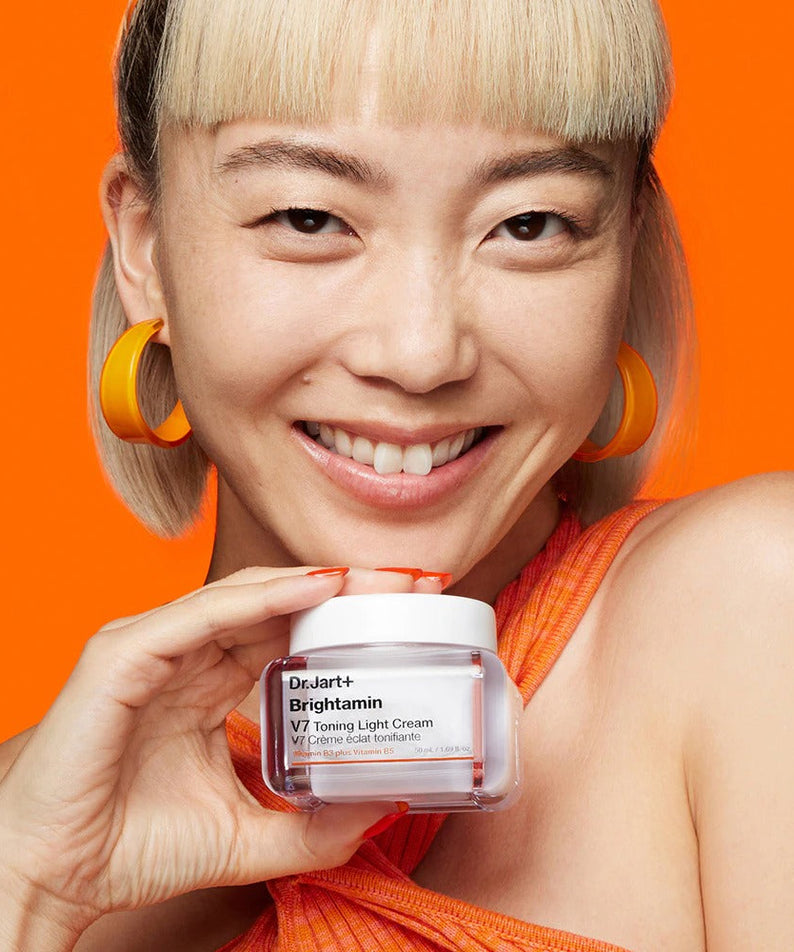 Woman smiling holding DR.JART+ Brightamine V7 Toning Light Cream against orange background