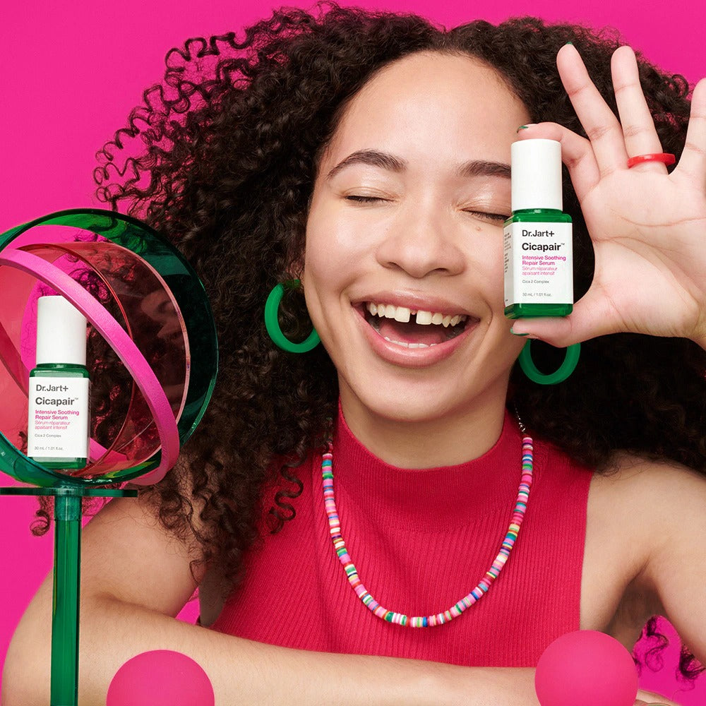 Smiling woman holding DR.JART+ CicapairI ntensive Soothing Repair Serum 30ml against pink background.