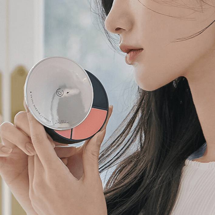 Woman holding DR.ALTHEA Swing Mirror Cushion (Cushion+Lip+Cheek) 15g near face, showcasing compact design for versatile makeup use.