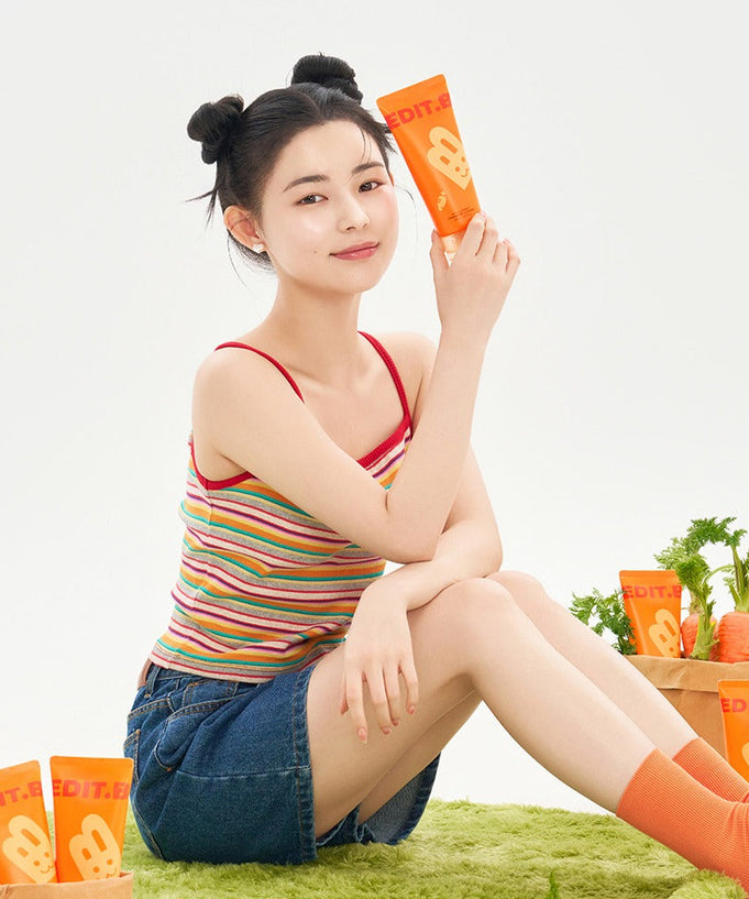 Person holding EDIT.B Joyful Carrot Foaming Gel Cleanser 150ml with orange packaging, surrounded by product tubes and fresh carrots.