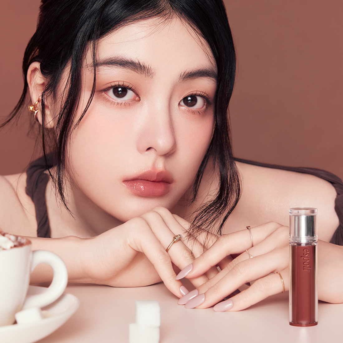 Woman wearing ESPOIR Couture Lip Tint Glaze [NEW COLOR], displayed with lip tint bottle and a cup of coffee in the foreground.