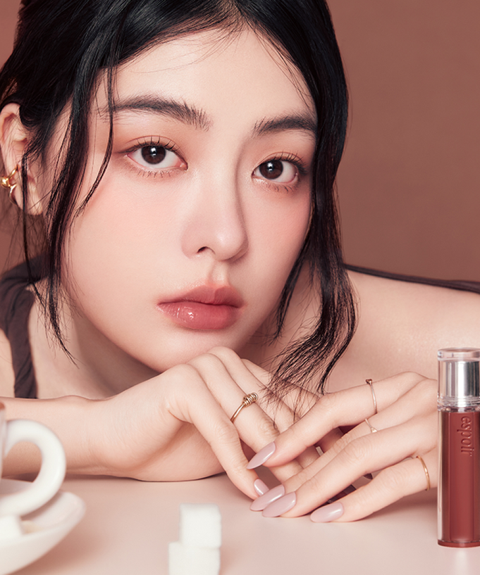 Woman wearing ESPOIR Couture Lip Tint Glaze [NEW COLOR], displayed with lip tint bottle and a cup of coffee in the foreground.