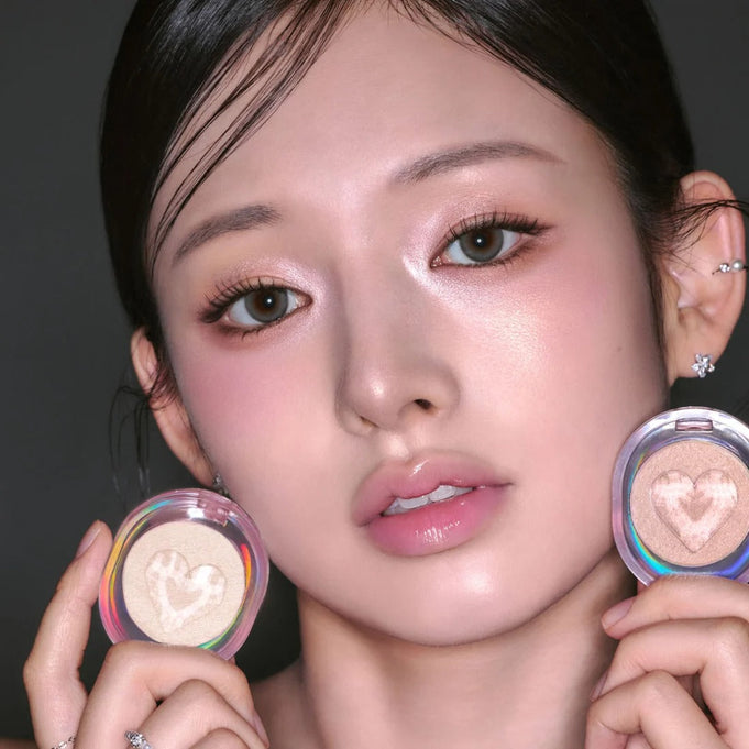 Woman holding ETUDE Shooting Beam Highlighter 3g with heart design, showing shimmering glow on cheekbones.