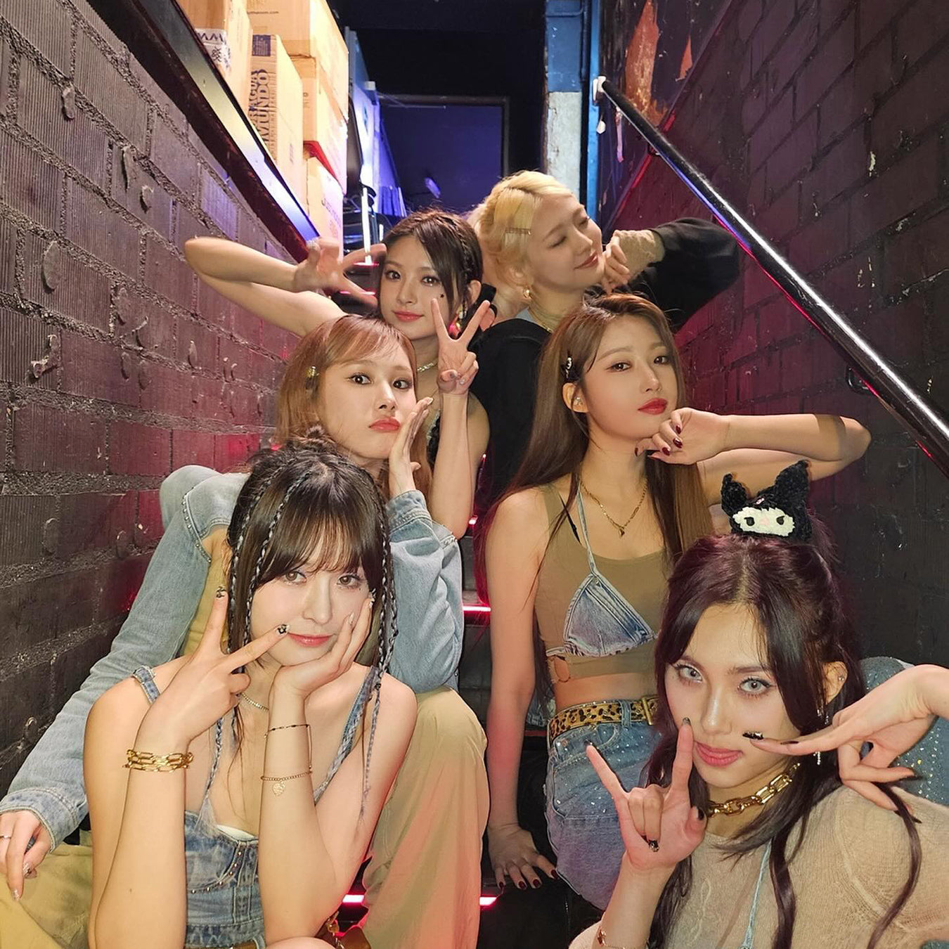 Six people posing playfully on stairs, with brick walls and colorful lighting.